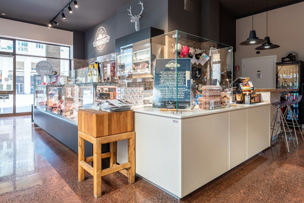 Macelleria con bancone frigo in vetro, sgabello di legno e pareti grigio scuro.