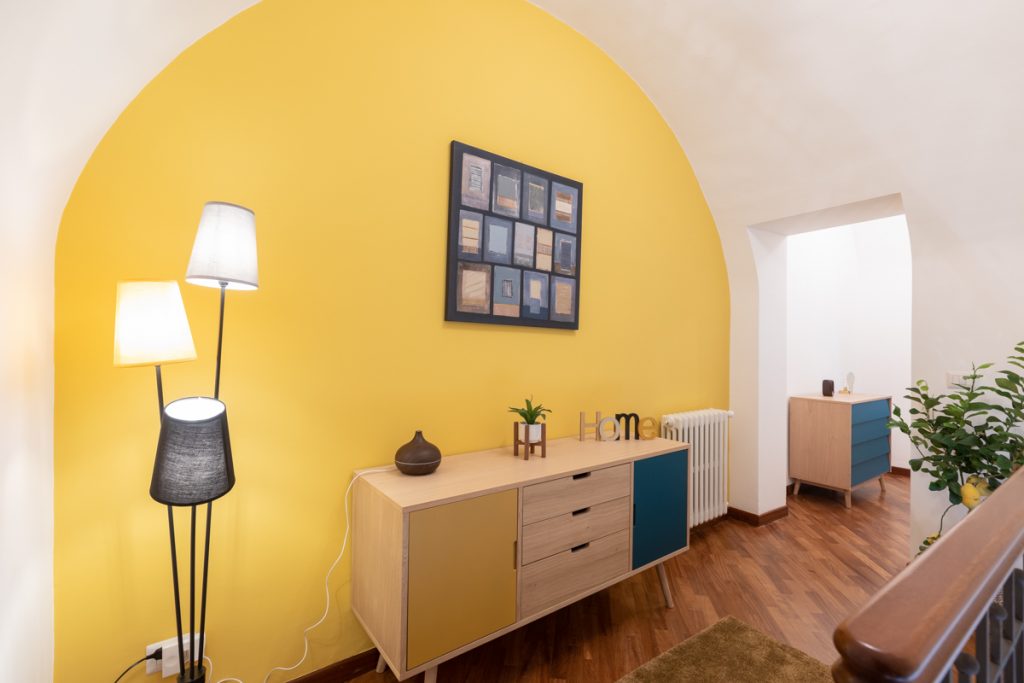 Corridoio con arco giallo dipinto sulla parete. Quadro appeso alla parete sopra un mobile in legno giallo e blu.