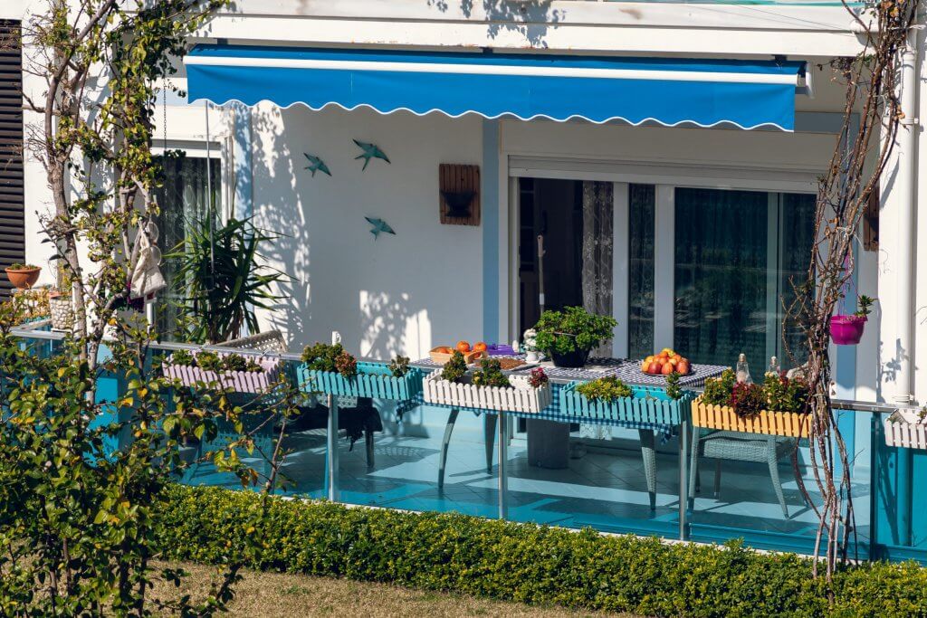 Balcone con fiori in vaso, finestra laterale bianco, tende da sole blu