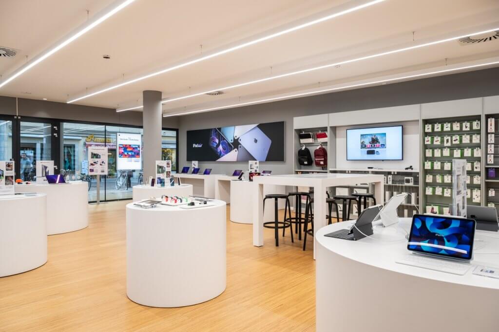 Store Apple con sgabelli neri, tavoli bianchi, computer, accessori, telefoni e tablet accesi con colori sgargianti