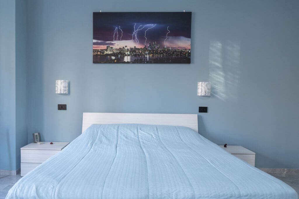 Camera da letto con applique da parete, quadro sulla testiera del letto, lenzuola e pareti azzurro pastello